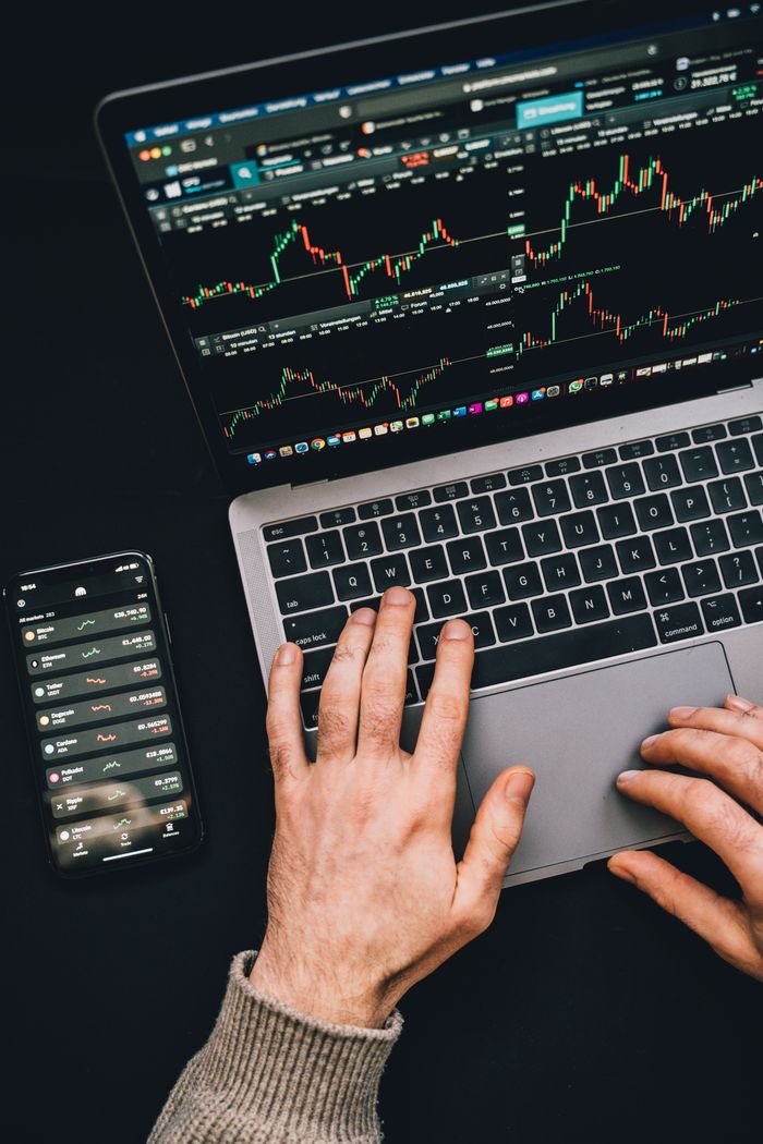 Des mains tapent sur un ordinateur portable avec des graphiques de cryptomonnaies et l’écran d’un smartphone visible, au centre de la finance numérique.