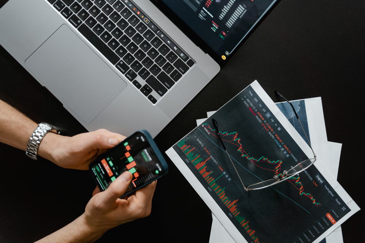 contact-img Hands using smartphone beside laptop with stock charts, showcasing digital trading.